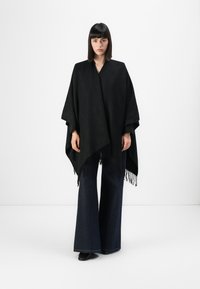 Zwarte poncho van textuurstof, met een V-hals ontwerp en franjerand. Gecombineerd met brede, donkere denim broek.