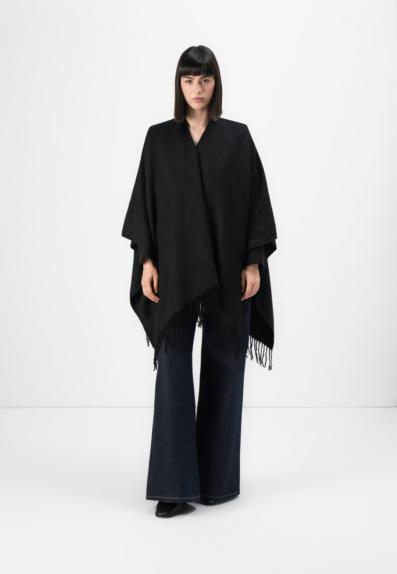 Zwarte poncho van textuurstof, met een V-hals ontwerp en franjerand. Gecombineerd met brede, donkere denim broek.