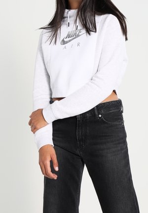 Žena v bílém krátkém mikině Nike Air a černých džínách s vysokým pasem, stojící se založenýma rukama vepředu.