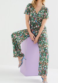 Květinový overal s krátkými rukávy, zeleným podkladem a barevnými květinovými vzory. Vázání v pase a široké, volné nohavice. Modré sandály na podpatku.