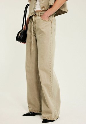 Osoba nosząca beżowe szerokie spodnie z troczkiem, krótka kurtka, trzyma małą czarną torebkę i nosi czarne czubate buty.