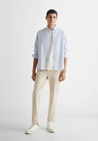 Lichtblauw overhemd met opgerolde mouwen, witte T-shirt eronder, crème broek en witte sneakers. Het overhemd heeft een klassieke pasvorm.