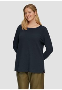 Lachende vrouw met lang blond haar, gekleed in een gestructureerde zwarte blouse met lange mouwen en olijfgroene broek, tegen een effen lichte achtergrond.