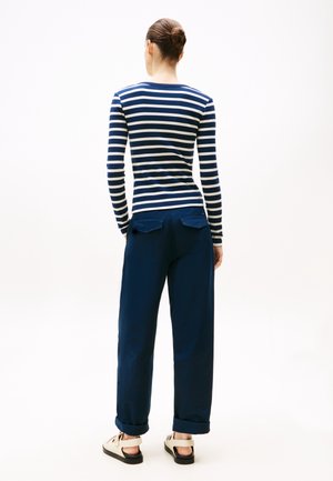 Femme aux cheveux bruns en chignon porte une chemise à manches longues rayée bleu marine et blanc, un pantalon bleu marine avec des revers et des sandales blanches, dos tourné.