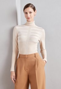 Beige Rollkragenpullover mit gerippter Textur und horizontalen Streifen. Kombiniert mit hoch taillierten, maßgeschneiderten braunen Hosen.