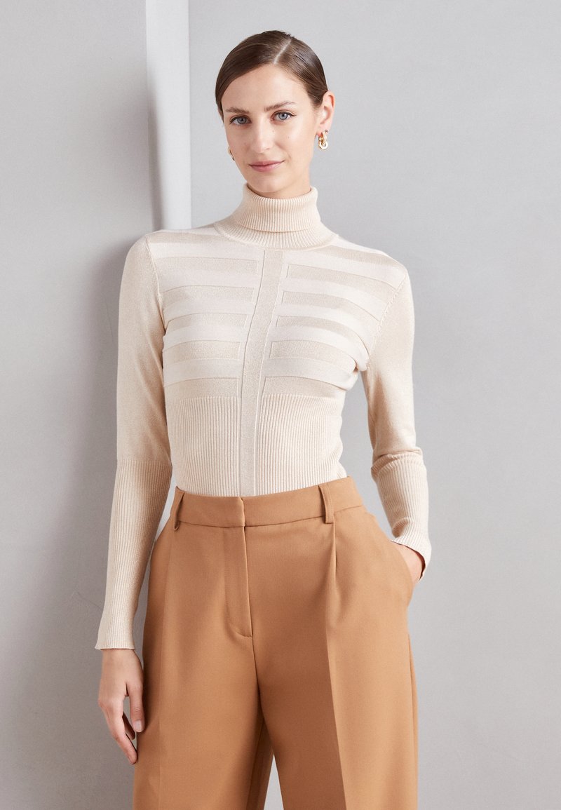 Beige Rollkragenpullover mit gerippter Textur und horizontalen Streifen. Kombiniert mit hoch taillierten, maßgeschneiderten braunen Hosen.