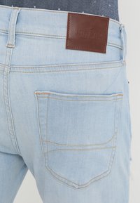 Jeans de mezclilla azul claro con una etiqueta de cuero marrón en la cintura, detalles en el bolsillo trasero y costuras a contraste.