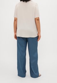 Persona che indossa una blusa beige chiaro a maniche corte e pantaloni blu larghi, in piedi con le spalle rivolte verso di noi su uno sfondo neutro.