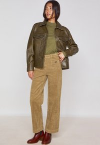 Giacca di pelle marrone, top verde oliva, pantaloni in corduroy beige a gamba larga e stivaletti marroni. Design semplice e pulito con dettagli di cucitura visibili.