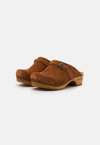 Zoccoli in suede marrone con suola in legno, apertura ampia e una fascia metallica decorativa. La texture è morbida e presenta un design a punta arrotondata.