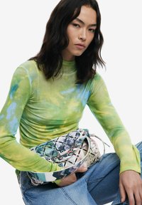 Grünes und blaues Batik-Hemd mit langen Ärmeln, kombiniert mit einer gemusterten Tasche in verschiedenen Farben und mit Metallakzenten, getragen zu blauen Denim-Jeans.