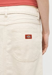 Vedere din spate a unei persoane care poartă pantaloni Dickies de culoare crem, cu buzunarul din spate vizibil și cu o etichetă roșie cu logo-ul Dickies sub talie.