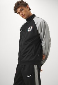 Μαύρο αθλητικό τζάκετ με γκρίζες μανίκες, φερμουάρ, πλαϊνές τσέπες και στρογγυλό λογότυπο που περιλαμβάνει ένα 'B'. Συνδυασμένο με αντίστοιχες παντελόνες.