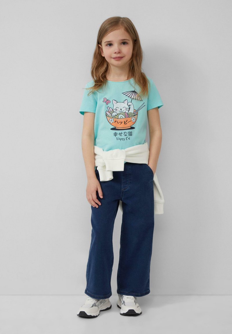 T-shirt bleu clair avec un motif de chat dans un bol, associé à un pantalon en denim large bleu foncé et des baskets blanches. Un pull est noué autour de la taille.