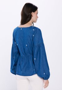 Denimbluse mit langen, puffigen Ärmeln. Blauer Stoff mit weißen gestickten Akzenten. Die Rückenform ist leicht gerafft für einen entspannten Sitz.