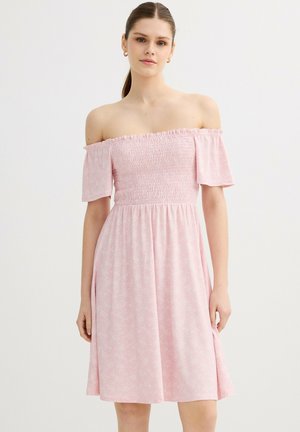 Joven mujer con un vestido rosa claro sin hombros, con manga corta y cuerpo fruncido, de pie frente a un fondo liso y claro.