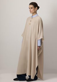 Beige ponšo pehmest kangast, millel on vabalt rippuv disain, külgmised lõhikud ja kolm silmatorkavat kuldset nööpi ees.