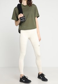 Camisa de manga corta de malla verde oliva, leggings crema con textura y patrones de ventilación, zapatillas deportivas negras y una toalla negra.