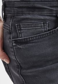 Jean en denim noir avec une vue rapprochée de la poche arrière, mettant en avant des détails de couture et une texture lisse. Main placée près de la poche.