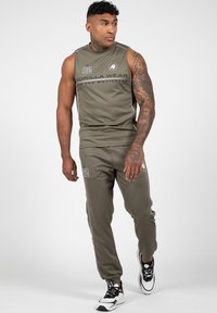 Atletische outfit in olijfgroen; mouwloos shirt met logo en nummer 82, bijpassende joggers met zijstrepen en logo; gecombineerd met zwart-witte sneakers.