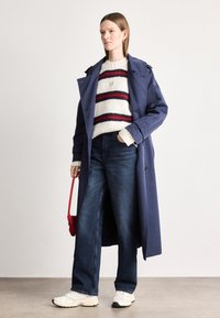 Cămașă lungă de marina, pulover gros tricotat cu dungi roșii și negre, blugi evazați albaștri, adidași albi și o geantă de umăr mică roșie.