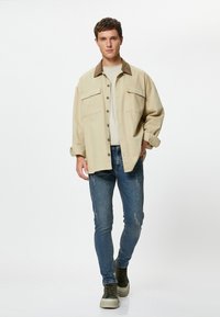 Modelo masculino caminando, vistiendo una camisa chaqueta oversized beige, camiseta ligera beige, jeans ajustados azules y zapatillas oscuras verdes, contra un fondo blanco.