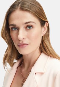 Lys pink blazer med slagkraver, tekstureret stof og en guldkædehalssmykke med en vedhæng. Modellen har bølget, mellemlangt brunt hår.