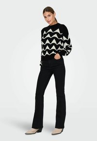 Maglione a maglia con un motivo a zigzag bianco e nero, abbinato a jeans neri a zampa d'elefante e scarpe con tacco beige. Tessuto strutturato, scollatura rotonda.