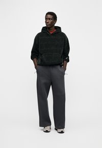 Μαύρο φούτερ με κουκούλα από υφασμένο ύφασμα με επαναλαμβανόμενο μοτίβο λογότυπου, σε συνδυασμό με χαλαρά σκούρα γκρι παντελόνια και μπεζ αθλητικά παπούτσια.