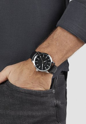 Schwarze Armbanduhr mit strukturiertem schwarzem Zifferblatt, silbernen Stundenmarkierungen und schwarzem Lederarmband, getragen an einem Handgelenk mit grauem Hemd.