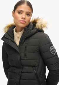 Chaqueta de plumas negra con capucha con ribete de piel, patrón de acolchado horizontal, bolsillos con cremallera y parche de logo en la manga.