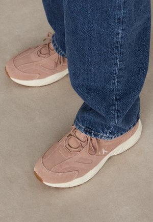Füße mit pinken Sneakern und blauen Jeans, stehend auf einem beigen Boden.