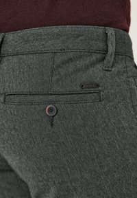 Nahaufnahme einer Tasche aus dunkelgrauer, strukturierter Hose mit Knopf und kleinem schwarzem Markenetikett, getragen mit einem dunkelroten Oberteil.