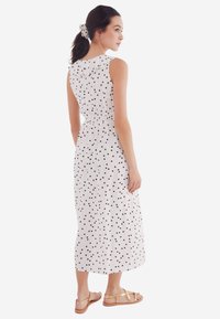 Witte mouwloze jurk met zwarte stippen, gerimpelde taille en een knielange zoom. Gestyled met een bijpassende haarband en platte sandalen.