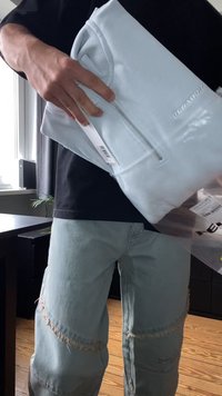 Felpa azzurra con zip realizzata in tessuto morbido, dotata di colletto a coste e logo impresso. Imballaggio in plastica trasparente visibile nella mano.