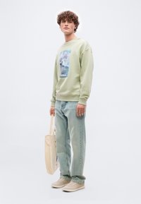 Νεαρός άνδρας με σγουρά μαλλιά, φοράει ανοιχτό πράσινο φούτερ με σχέδιο, ανοιχτό μπλε τζιν, μπεζ αθλητικά παπούτσια και κρατάει μια μπεζ τσάντα ώμου.