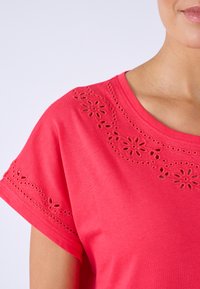 Mujer con una camiseta roja de manga corta con patrones de recortes florales bordados en el escote y las mangas.