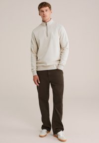 Felpa leggera beige chiaro con zip a metà, maniche a costine, abbinata a pantaloni dritti marrone scuro e sneakers bianche con dettagli beige.