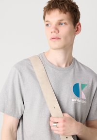 Grå bomulds T-shirt med et lille farverigt Calvin Klein-logo. Lysefarvet rem over skulderen og en sølvring på hånden.