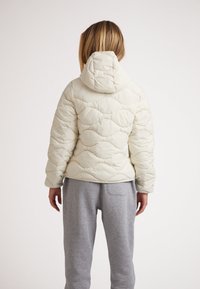 Puffig vit jacka med huva, med en quiltad textur och vågiga mönster. Bärs över grå mjukisbyxor. Synliga sömdetaljer.