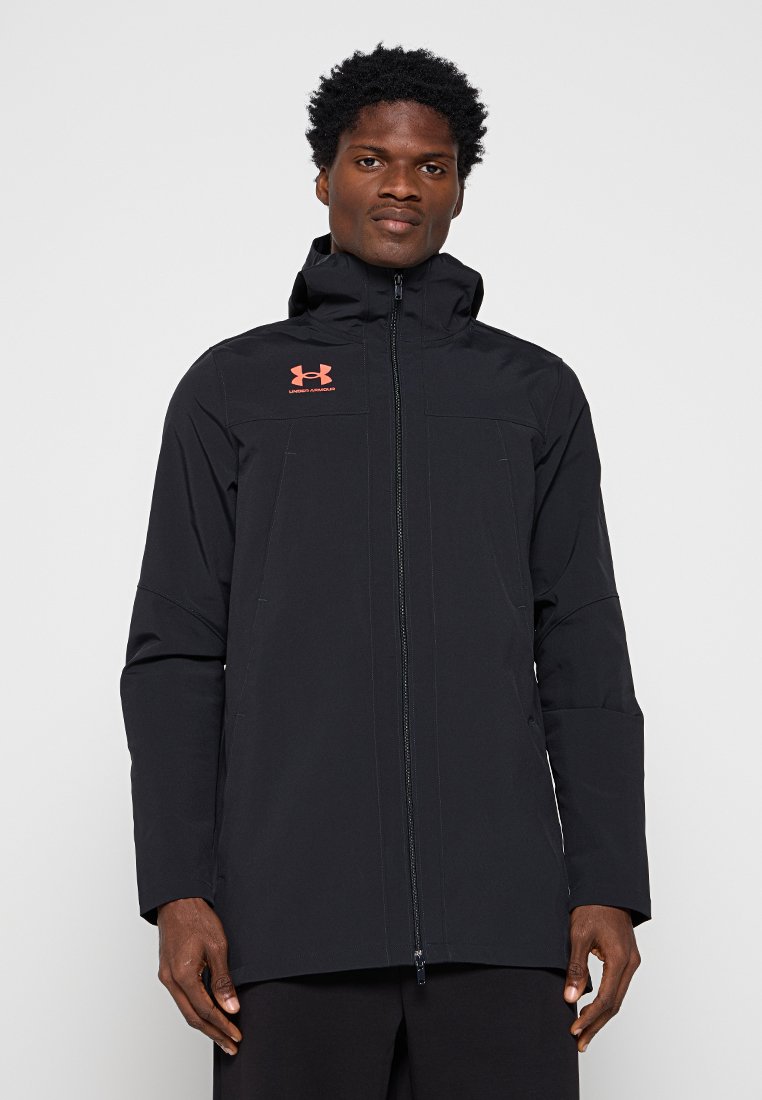 Veste imperméable noire avec un col haut et une fermeture éclair pleine sur le devant. Présente un logo sur la poitrine et des poches latérales. Matériau lisse et léger.
