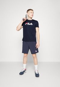 Ναυτικό μπλε φανέλα Fila με άσπρο και κόκκινο λογότυπο, συνδυασμένη με σκούρες γκρίζες βερμούδες που έχουν ένα μικρό λογότυπο, και αθλητικά παπούτσια με κόκκινες λεπτομέρειες.