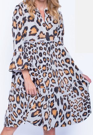 Kvinde iført en knælængde kjole med brun og sort leopardprint, lange ærmer og en samlet skørt, der holder nederkanten med den ene hånd.