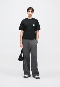 Μαύρο βαμβακερό μπλουζάκι με τσέπη στο στήθος, σε συνδυασμό με γκρι φόρμα. Το μοντέλο κρατά μια μικρή μαύρη τσάντα και φοράει γκρι αθλητικά παπούτσια.