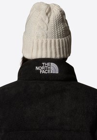 Czarna polarowa kurtka z wysokim kołnierzem z haftowanym białym logo. Na głowie noszona jest beanie w odcieniu kremowym z warkoczowym splotem.