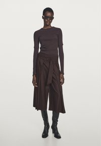 Femme portant un haut à manches longues marron foncé, des culottes larges ceinturées, des bottes noires jusqu'aux genoux et des lunettes de soleil, se tenant devant un fond clair. 