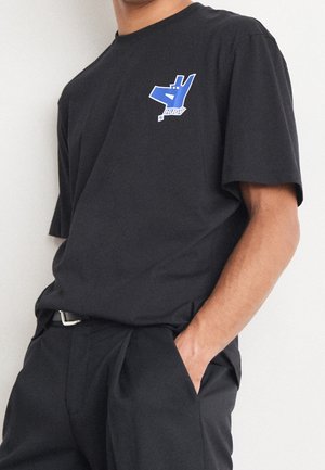 Personne portant un t-shirt noir avec un petit motif bleu sur le côté gauche de la poitrine et un pantalon noir, une main dans la poche.