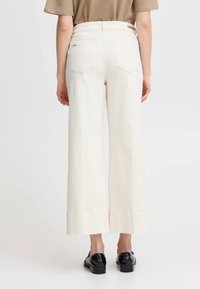 Pantaloni in denim a gamba larga di colore crema con due tasche posteriori e orlo piegato. Indossati con scarpe nere, mostrando una texture del tessuto liscia.