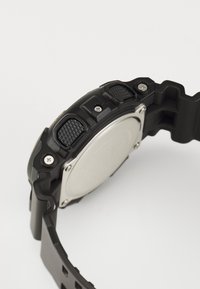 Montre noire avec un bracelet en caoutchouc texturé, des accents en métal et un boîtier rond. Caractéristiques de boutons proéminents et de vis visibles sur le côté.