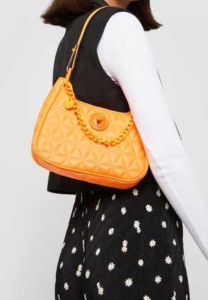 Vrouw draagt een mouwloos zwart vest en zwarte broek met madeliefjespatroon, en draagt een gequilte feloranje handtas met kettingdetail over de schouder.